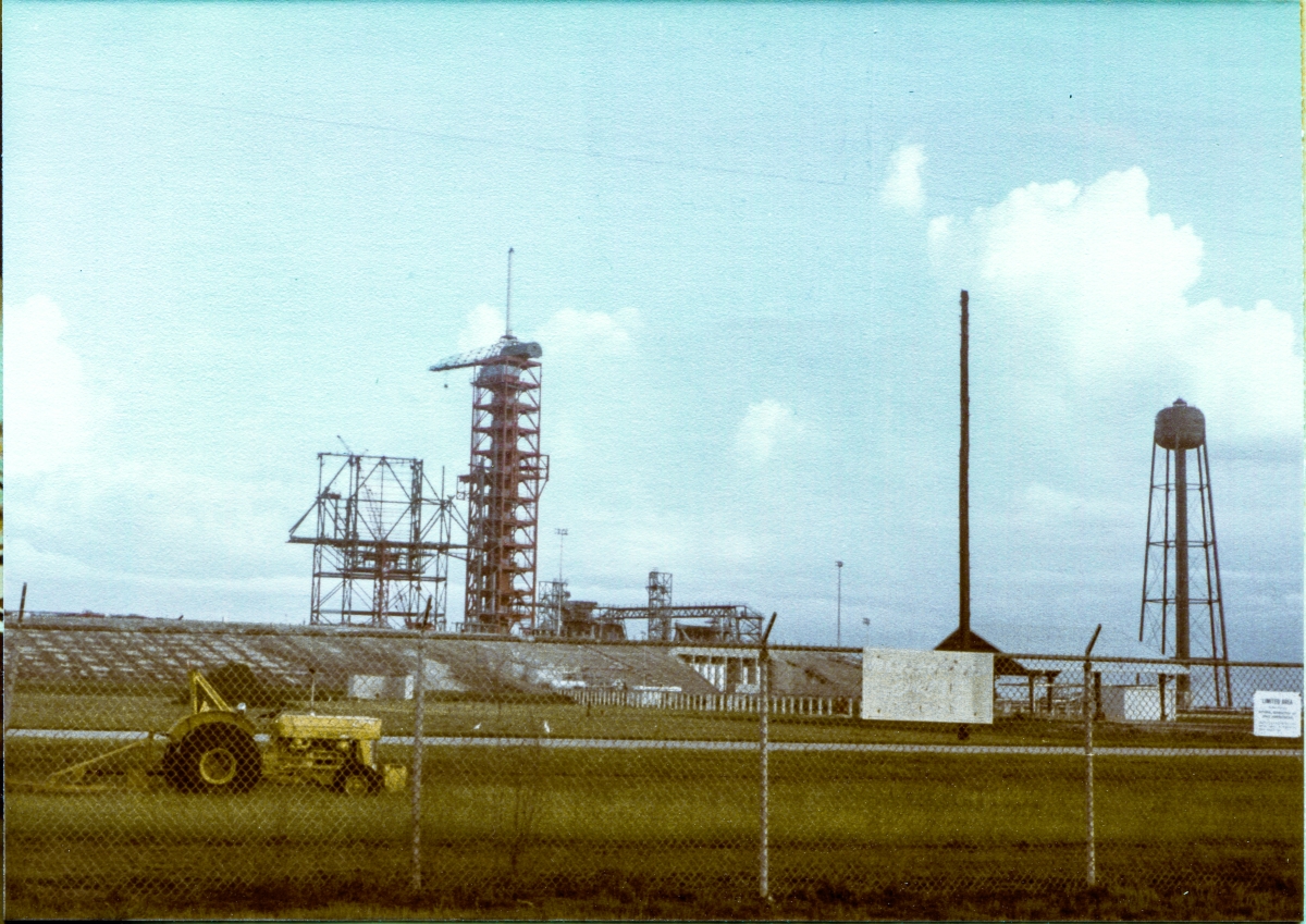 Image 004. Viewed from the turn in the road where the pad access road from A1A meets the pad perimeter, just outside the perimeter fence, the growing steel skeleton of the RSS stands in sharp relief against the sky, supported by the temporary falsework framing upon which it is resting. In this generalized view of Space Shuttle Launch Pad 39-B at the Kennedy Space Center, Florida, you can see the fifty-foot high concrete mass of the pad itself, and more or less from left to right, the RSS upon its falsework, the tall red multi-platformed framing of the FSS, the 9099 Building, the MLP Access Stair Tower, the North Piping Bridge, with the High Pressure Gas Area cutout into the pad slope beneath it, and beneath that, the supports for the cross-country piping run leading to the Hypergol Oxidizer (N2O4) Storage Facility which is partially-obscured by signage on the pad perimeter fence. At far right stands the SSW Water Tower. Photo by James MacLaren. Image 004. Viewed from the turn in the road where the pad access road from A1A meets the pad perimeter, just outside the perimeter fence, the growing steel skeleton of the RSS stands in sharp relief against the sky, supported by the temporary falsework framing upon which it is resting. In this generalized view of Space Shuttle Launch Pad 39-B at the Kennedy Space Center, Florida, you can see the fifty-foot high concrete mass of the pad itself, and more or less from left to right, the RSS upon its falsework, the tall red multi-platformed framing of the FSS, the 9099 Building, the MLP Access Stair Tower, the North Piping Bridge, with the High Pressure Gas Area cutout into the pad slope beneath it, and beneath that, the supports for the cross-country piping run leading to the Hypergol Oxidizer (N2O4) Storage Facility which is partially-obscured by signage on the pad perimeter fence. At far right stands the SSW Water Tower. Photo by James MacLaren.